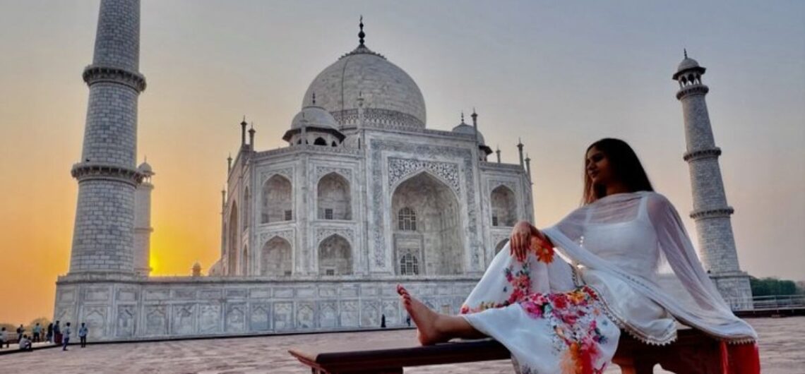 Woman in traditional white attire sitting in front of the Taj Mahal at sunrise, capturing the beauty of India's most famous UNESCO World Heritage Site.